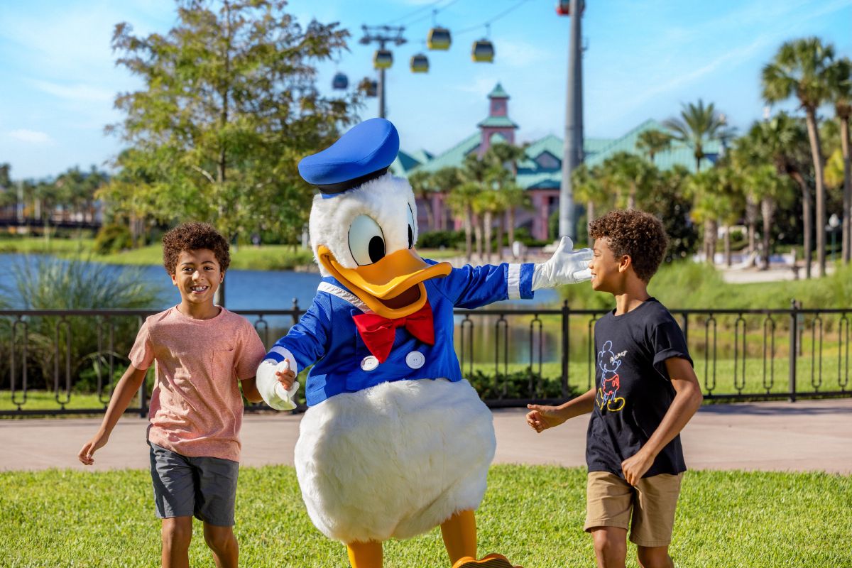 Two boys laughing and playing with Donald Duck on a green lawn at Disney's Caribbean Beach Resort, with the Disney Skyliner gondola and palm trees in the background