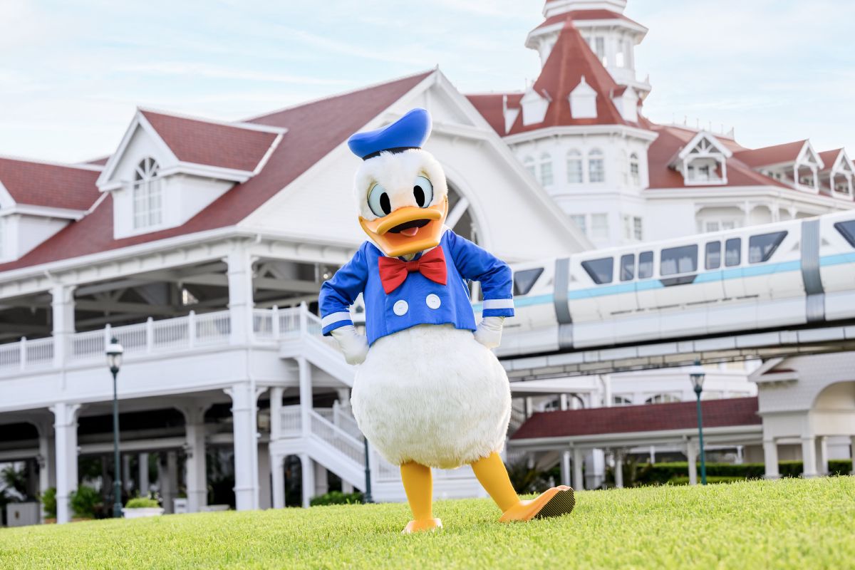 Donald Duck posing on the lawn in front of Disney's Grand Floridian Resort and Spa, with the monorail passing behind the white Victorian-style building