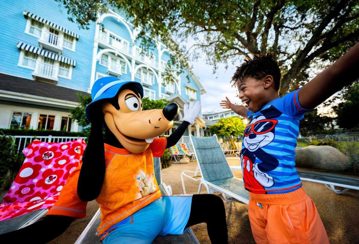 A child in a blue swim shirt high-fiving Goofy poolside at Disney's BoardWalk Inn, with the blue resort building in the background