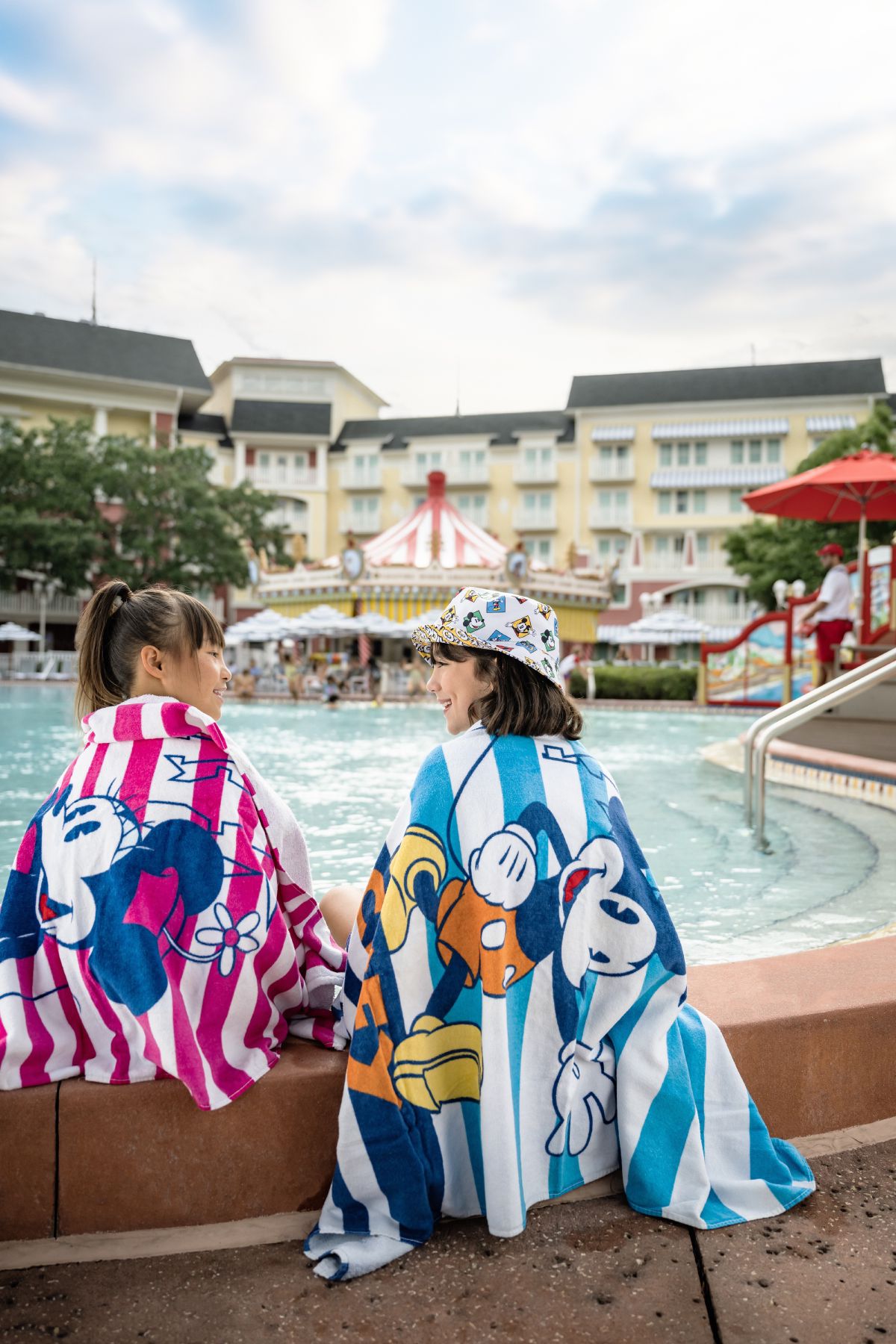 Two guests wrapped in colorful Disney character towels by the pool at Disney's BoardWalk Inn, enjoying the resort before their room is ready.