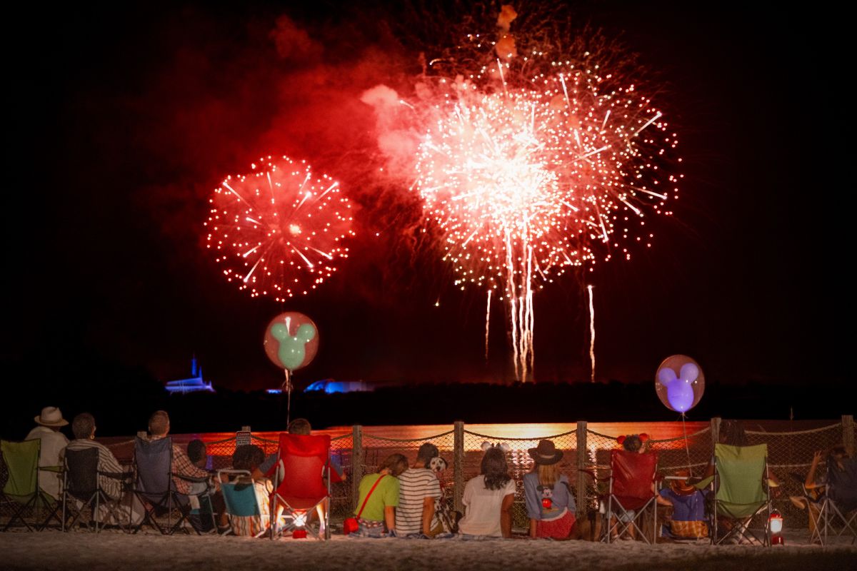 Guests watching Magic Kingdom Park fireworks from a waterfront viewing area at Walt Disney World Resort, with colorful bursts reflected on the lagoon.