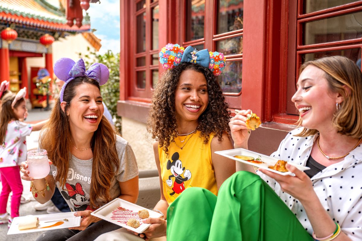Three friends sharing food and laughing together at a World Showcase pavilion during the EPCOT International Festival of the Arts.