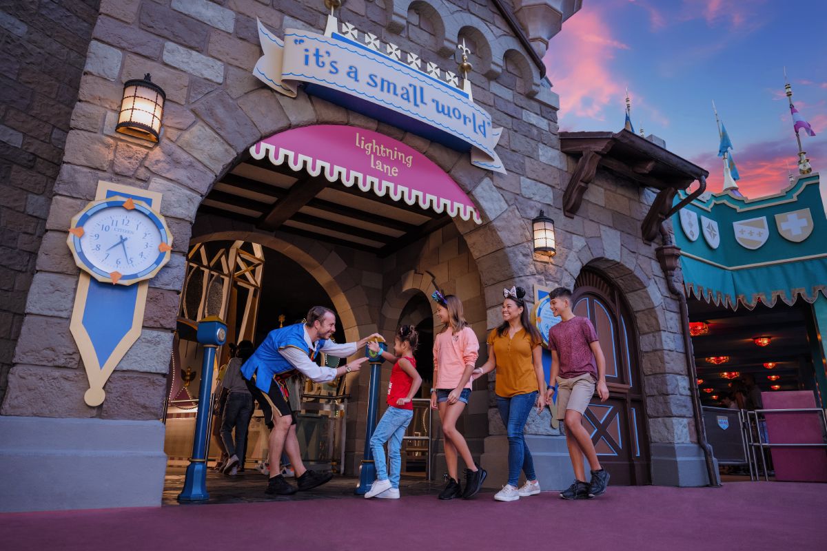 A Cast Member greets a young guest at the entrance of it’s a small world at Magic Kingdom Park. The family enters through the Lightning Lane under a fairytale-style archway with a themed clock and banners.