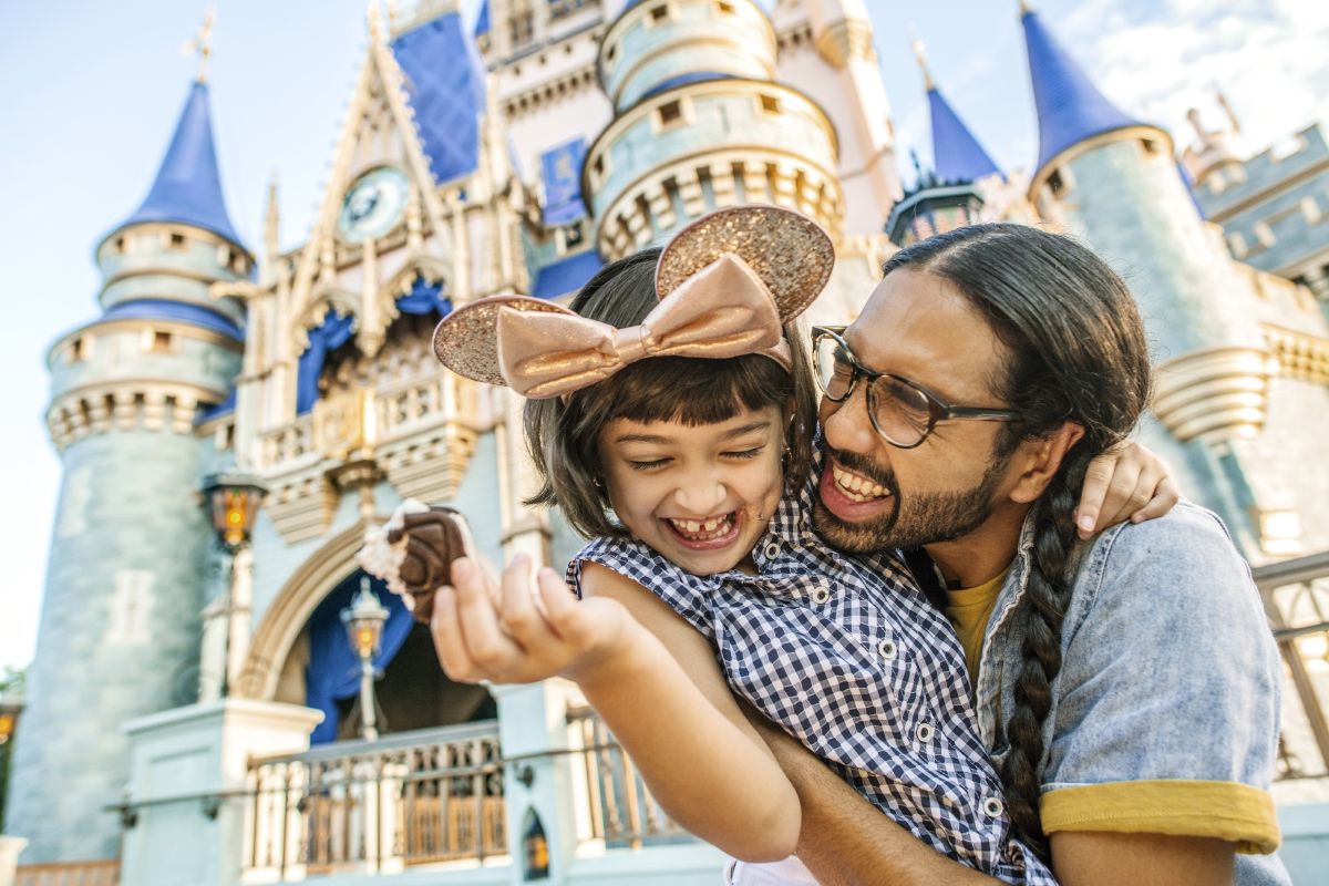 A father and young daughter wearing mouse ears share an ice cream treat in front of Cinderella Castle at Magic Kingdom Park, capturing the kind of memory families make on a Walt Disney World vacation.