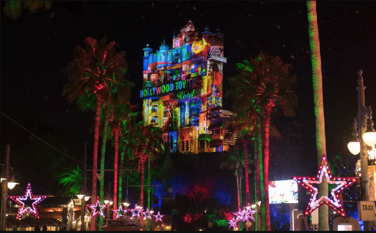 The Hollywood Tower Hotel covered in colorful holiday projections at night, framed by palm trees and lighted star decorations, part of the Christmas season at Walt Disney World.