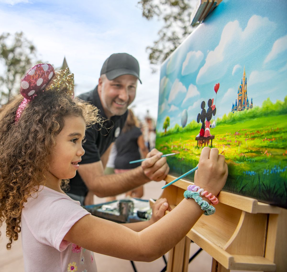 A father and daughter paint together at an outdoor easel during the EPCOT International Festival of the Arts, with a colorful Disney-themed mural in the background.