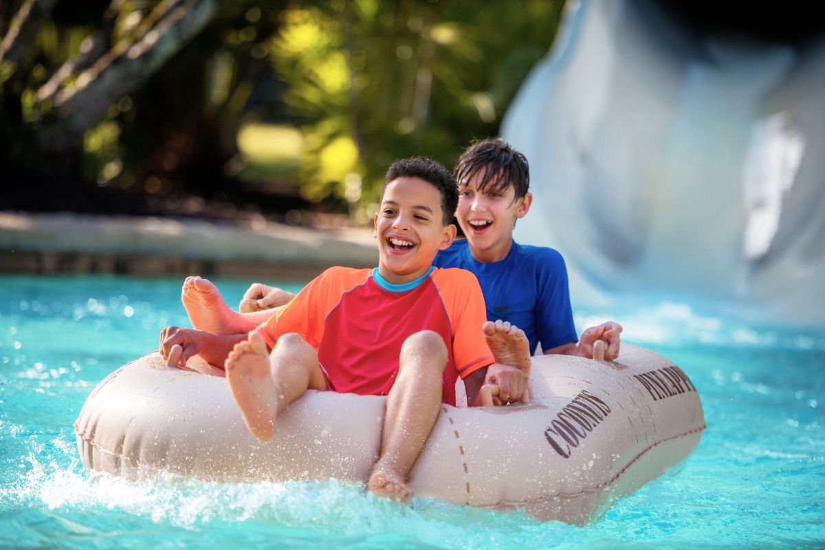 Two boys laughing on a raft as they speed down a water slide at Disney's Typhoon Lagoon Water Park, water spraying around them.