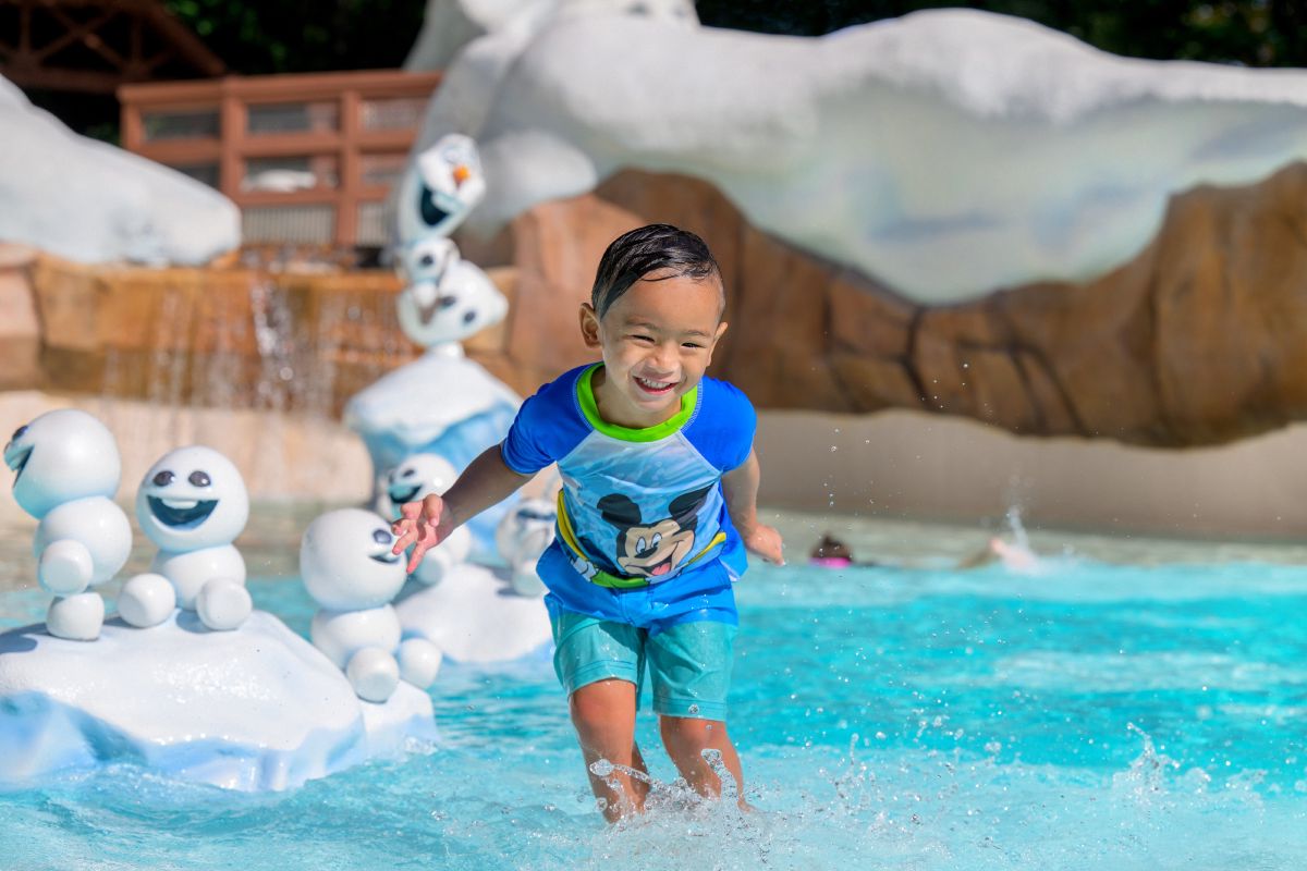 Young boy in a Mickey Mouse swimsuit grinning as he splashes through the Frozen-themed Tike's Peak play area at Disney's Blizzard Beach Water Park.