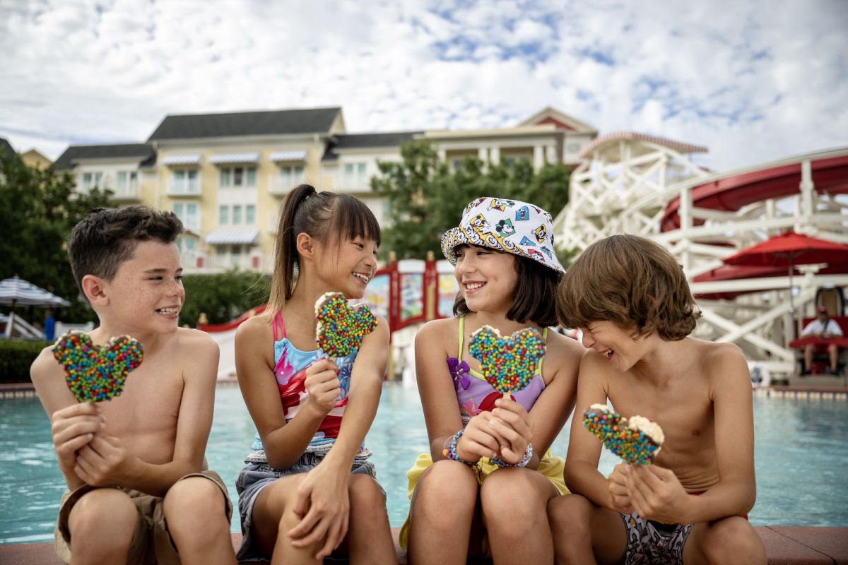 A group of kids laughing together at the pool at Disney's BoardWalk Inn, holding colorful drinks on a sunny day.